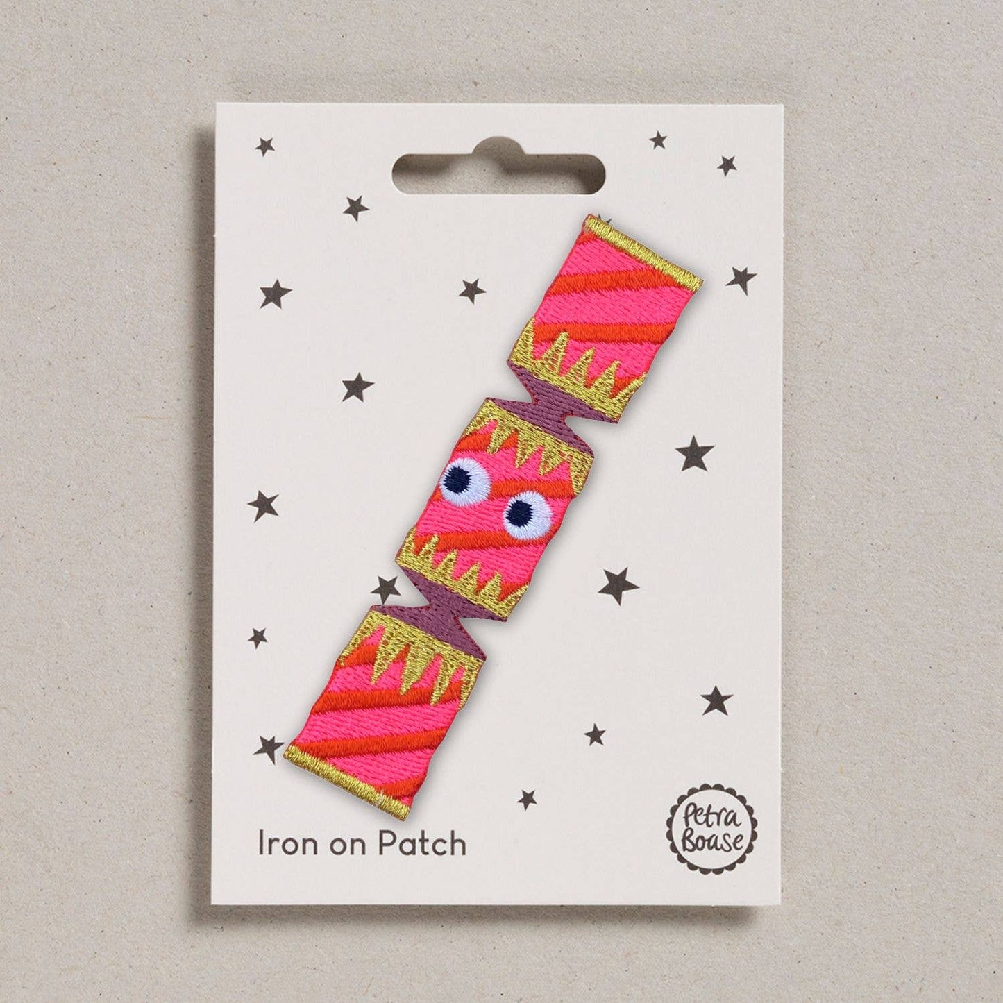 an embroidered Christmas cracker with goggly black eyes.It is pink in colour with purple and golden decoration on the wrapper. The background is white with black stars. 