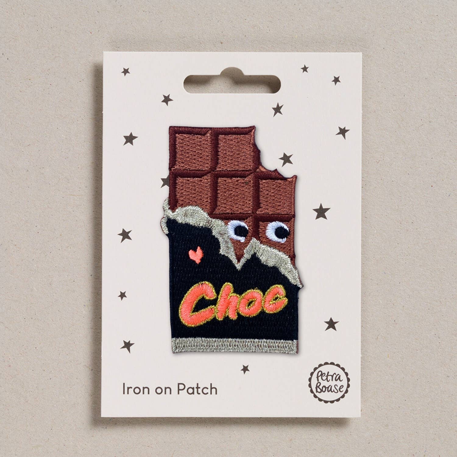 The Love Choc iron-on patch displayed in its original packaging, a cream-coloured card with scattered black stars. The Petra Boase branding is printed at the bottom, and "Iron on Patch" is written in simple text. The patch itself features a bitten chocolate bar with googly embroidered eyes, a torn wrapper, and the word "Choc" in a retro font.