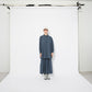 Model standing in front of a white photography backdrop, wearing the oversized blue checkered shirt and matching skirt. The relaxed silhouette is complemented by a casual cap and chunky trainers