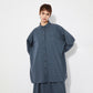 Model wearing the oversized blue checkered shirt with a matching skirt, standing with arms behind their back. The wide, draped silhouette of the shirt is highlighted, creating a structured yet relaxed effect