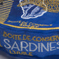 Close-up of a blue embroidered fabric item with gold and white text reading 'BOITE DE CONSERVE SARDINES L'HUILE' and a white fish logo.