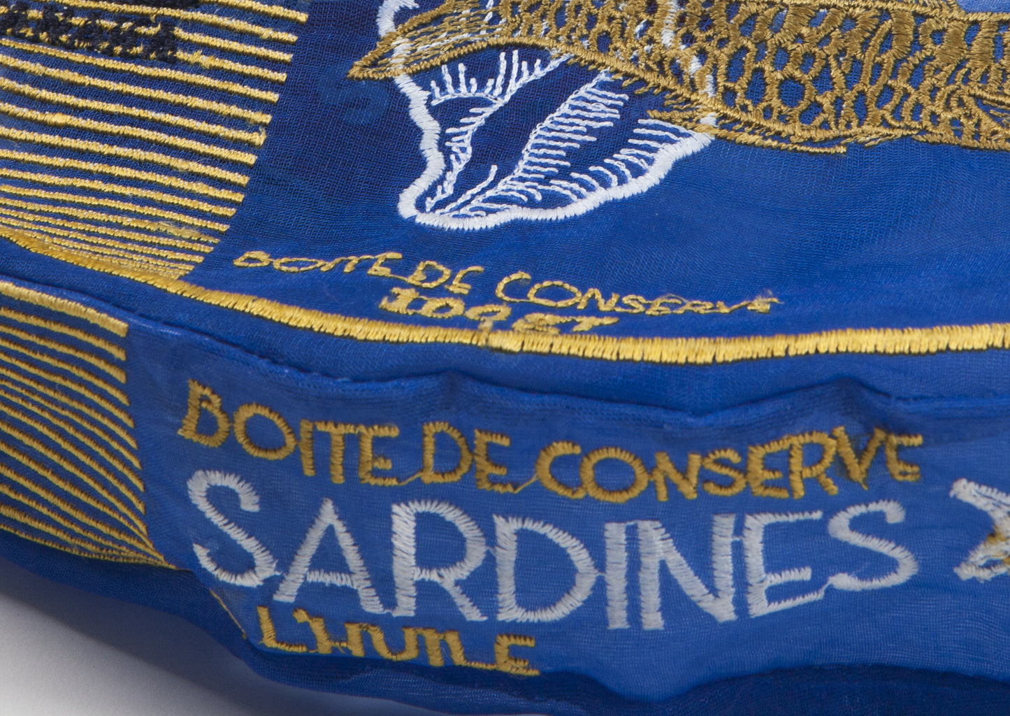 Close-up of a blue embroidered fabric item with gold and white text reading 'BOITE DE CONSERVE SARDINES L'HUILE' and a white fish logo.