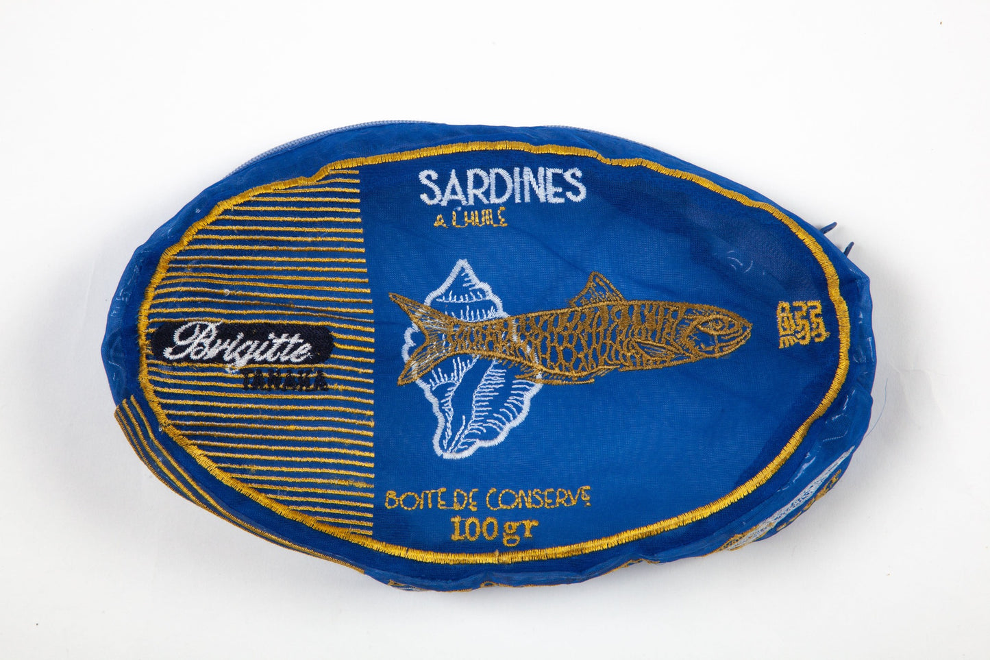 A single oval-shaped wrapped food product on a plain white background. The package is predominantly blue with gold/yellow border lines, white text reading 'SARDINES & LINGUES' and 'BOITE DE CASSERVE 100gr', a fish illustration, and a black oval label on the left that says 'Brigitte FINANCIA'.