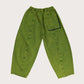 A pair of wide-leg, barrel-shaped trousers in a textured green shade with an elasticated waistband, side pockets, and a back patch pocket. The trousers feature displaced seams for a rounded leg shape, offering a modern and relaxed fit. Made from 20% recycled cotton and styled for unisex wear. The model wears a matching jacket with a black t-shirt underneath and striped socks with black shoes 