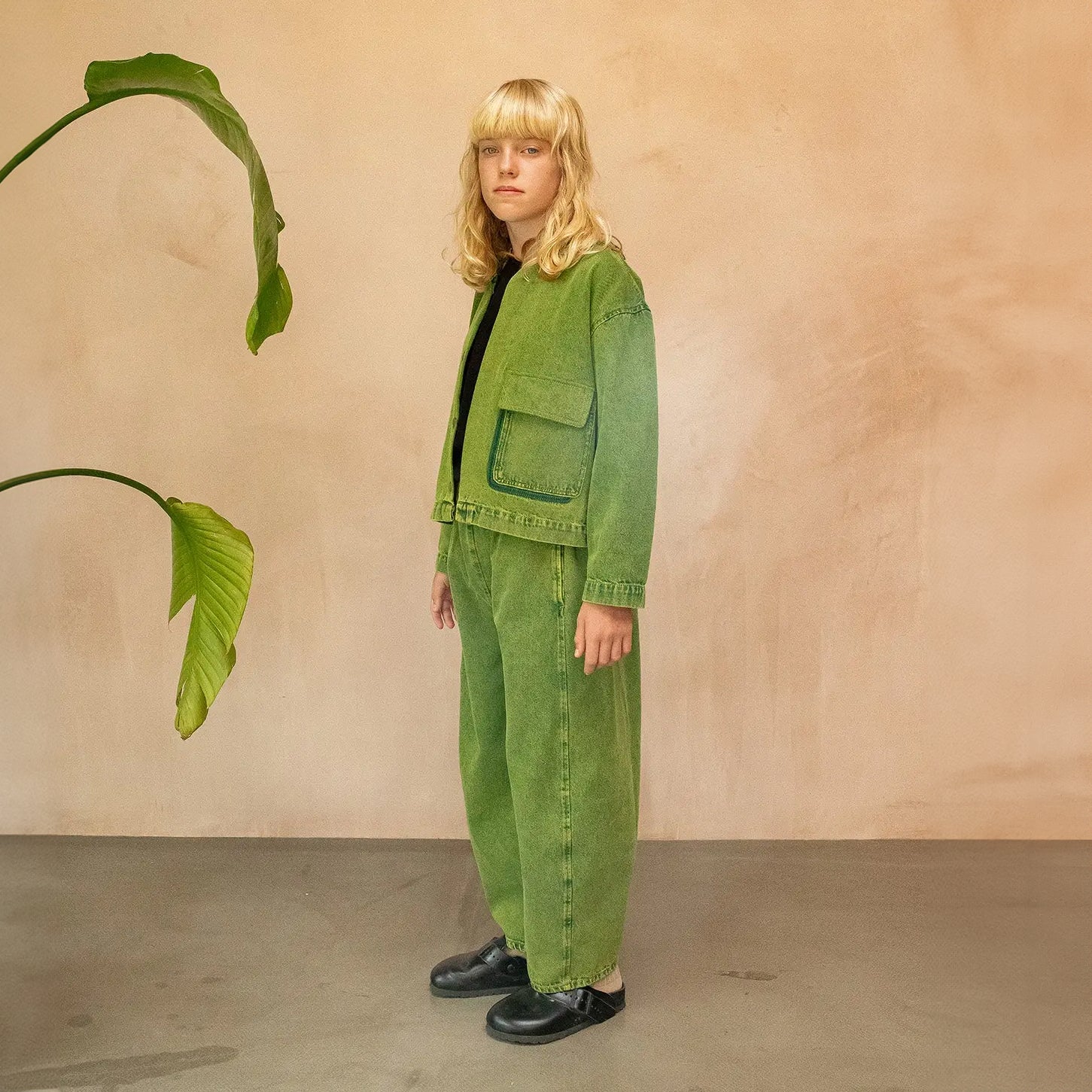 A pair of wide-leg, barrel-shaped trousers in a textured green shade with an elasticated waistband, side pockets, and a back patch pocket. The trousers feature displaced seams for a rounded leg shape, offering a modern and relaxed fit. Made from 20% recycled cotton and styled for unisex wear. The model wears a matching jacket with a black t-shirt underneath and striped socks with black mules