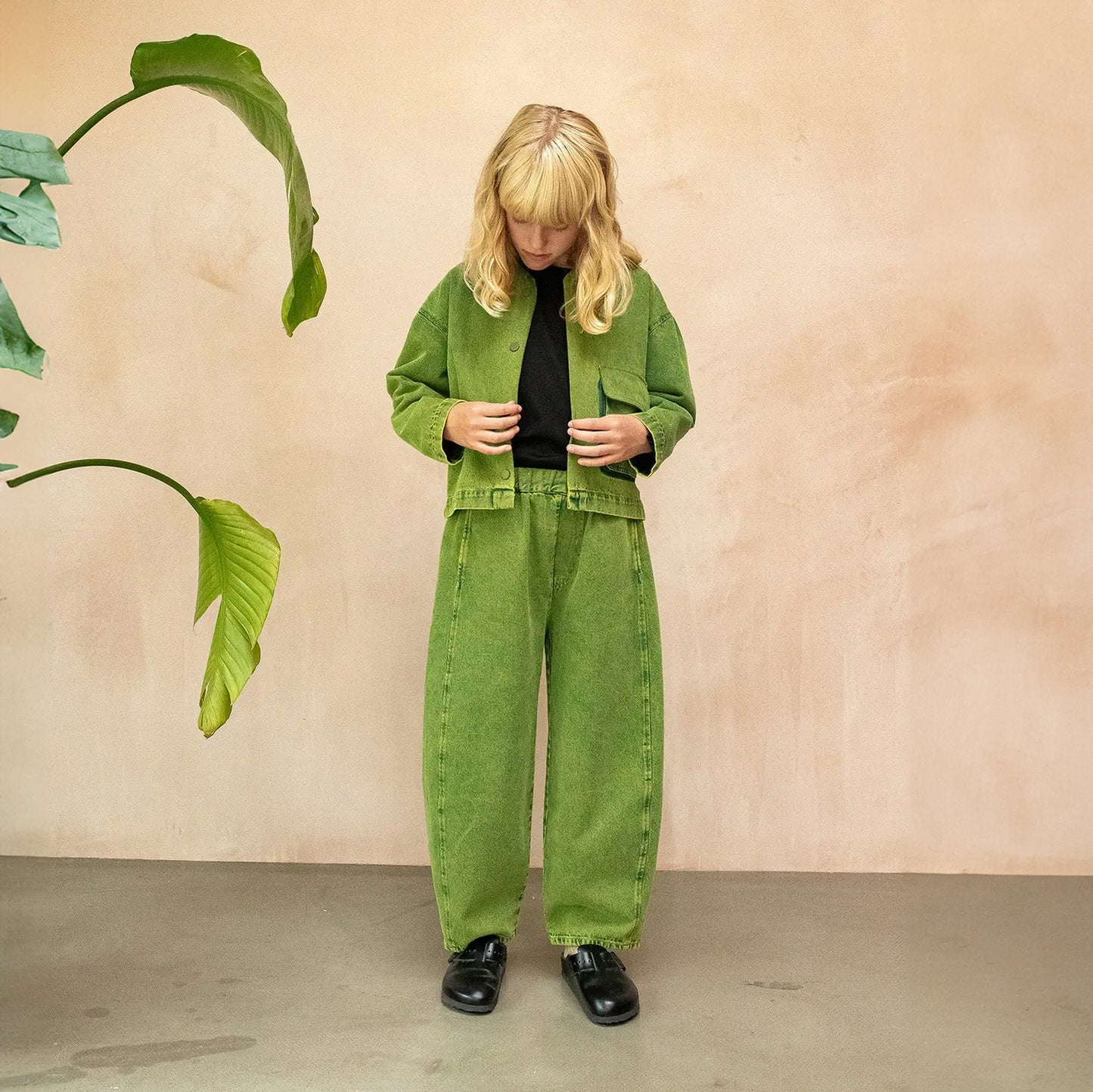 A pair of wide-leg, barrel-shaped trousers in a textured green shade with an elasticated waistband, side pockets, and a back patch pocket. The trousers feature displaced seams for a rounded leg shape, offering a modern and relaxed fit. Made from 20% recycled cotton and styled for unisex wear. The model wears a matching jacket with a black t-shirt underneath and striped socks with black mules. She faces the camera as if starting to button the jacket