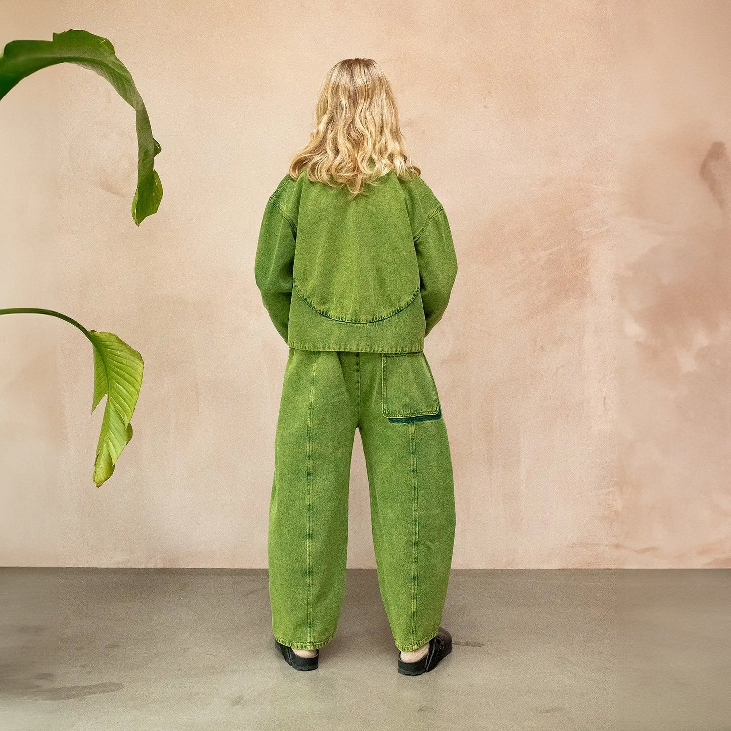 A pair of wide-leg, barrel-shaped trousers in a textured green shade with an elasticated waistband, side pockets, and a back patch pocket. The trousers feature displaced seams for a rounded leg shape, offering a modern and relaxed fit. Made from 20% recycled cotton and styled for unisex wear. The model wears a matching jacket with a black t-shirt underneath and striped socks with black mules. She is shown from the back