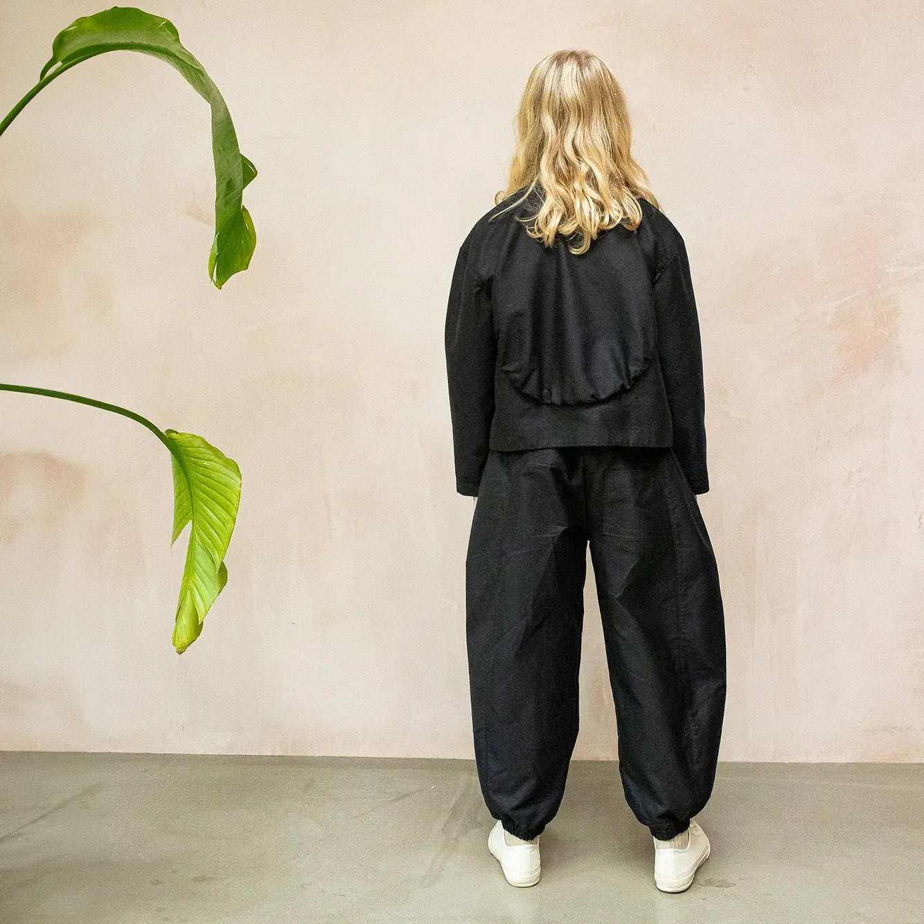 A pair of Otis Woven Wide-Leg Trousers in versatile black, showcasing a relaxed rounded silhouette, gathered cuffs, and an elastic waistband with an internal drawcord. The trousers are made from soft, washed double cotton and feature pockets for added practicality. The blonde haired model wears with a striped roll necked top and matching black jacket shown from the back