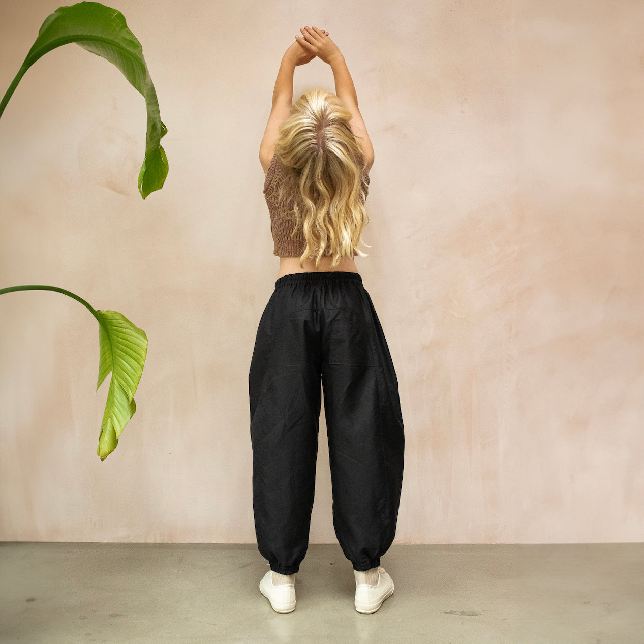 A pair of Otis Woven Wide-Leg Trousers in versatile black, showcasing a relaxed rounded silhouette, gathered cuffs, and an elastic waistband with an internal drawcord. The trousers are made from soft, washed double cotton and feature pockets for added practicality. The blonde haired model wears the Skye knitted vest from the same collection. She is shown from the back with her hands up in the air