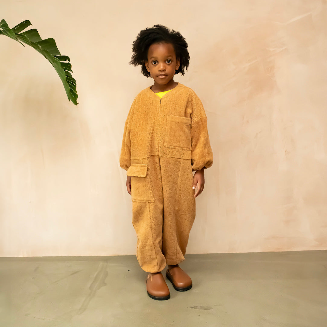 Studio Koter Rae Oversized Teddy Boilersuit in warm caramel, showcasing the soft texture of the 100% organic cotton teddy fleece. The image highlights the zip front, elasticated cuffs, and spacious patch pockets, emphasizing the cozy and functional design perfect for cooler days. The model wears a yellow t-shirt underneath and brown boots. She stands next to a banana leaf palm