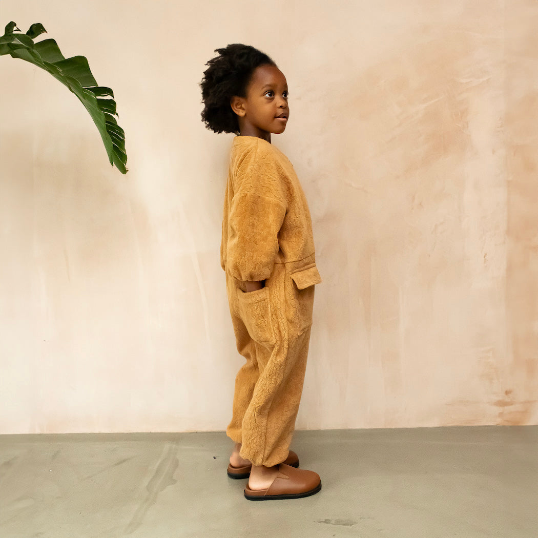 Studio Koter Rae Oversized Teddy Boilersuit in warm caramel, showcasing the soft texture of the 100% organic cotton teddy fleece. The image highlights the zip front, elasticated cuffs, and spacious patch pockets, emphasizing the cozy and functional design perfect for cooler days. The model wears a yellow t-shirt underneath and brown boots. She stands next to a banana leaf palm. The model is standing to the side