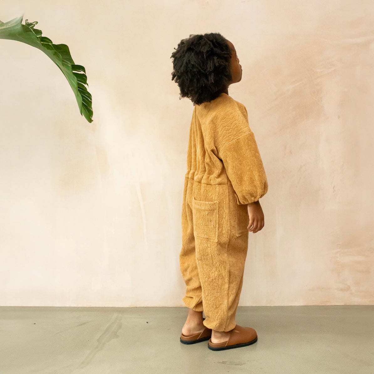 Studio Koter Rae Oversized Teddy Boilersuit in warm caramel, showcasing the soft texture of the 100% organic cotton teddy fleece. The image highlights the zip front, elasticated cuffs, and spacious patch pockets, emphasizing the cozy and functional design perfect for cooler days. The model wears a yellow t-shirt underneath and brown boots. She stands next to a banana leaf palm. The view is from the side of the jumpsuit