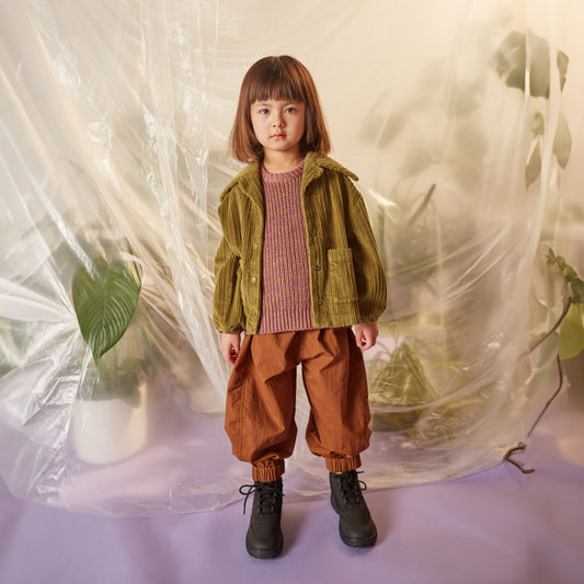 Studio Koter Skye Knitted Vest in a chunky half-cardigan stitch, featuring a boxy fit and round neckline. Made from soft cotton yarn in a mix of purple and olive green, creating a unique melange effect. The model wears it with an olive corduroy jacket, brown wide leg trousers, and lace up boots