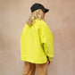 Close-up of a blonde, long-haired model wearing the Studio Koter River Teddy Sweatshirt in vibrant acid green, paired with beige trousers made from matching teddy fabric. The outfit is complemented by black trainers and a black cap, showcasing a stylish and comfortable look. The sweatshirt is shown from the back