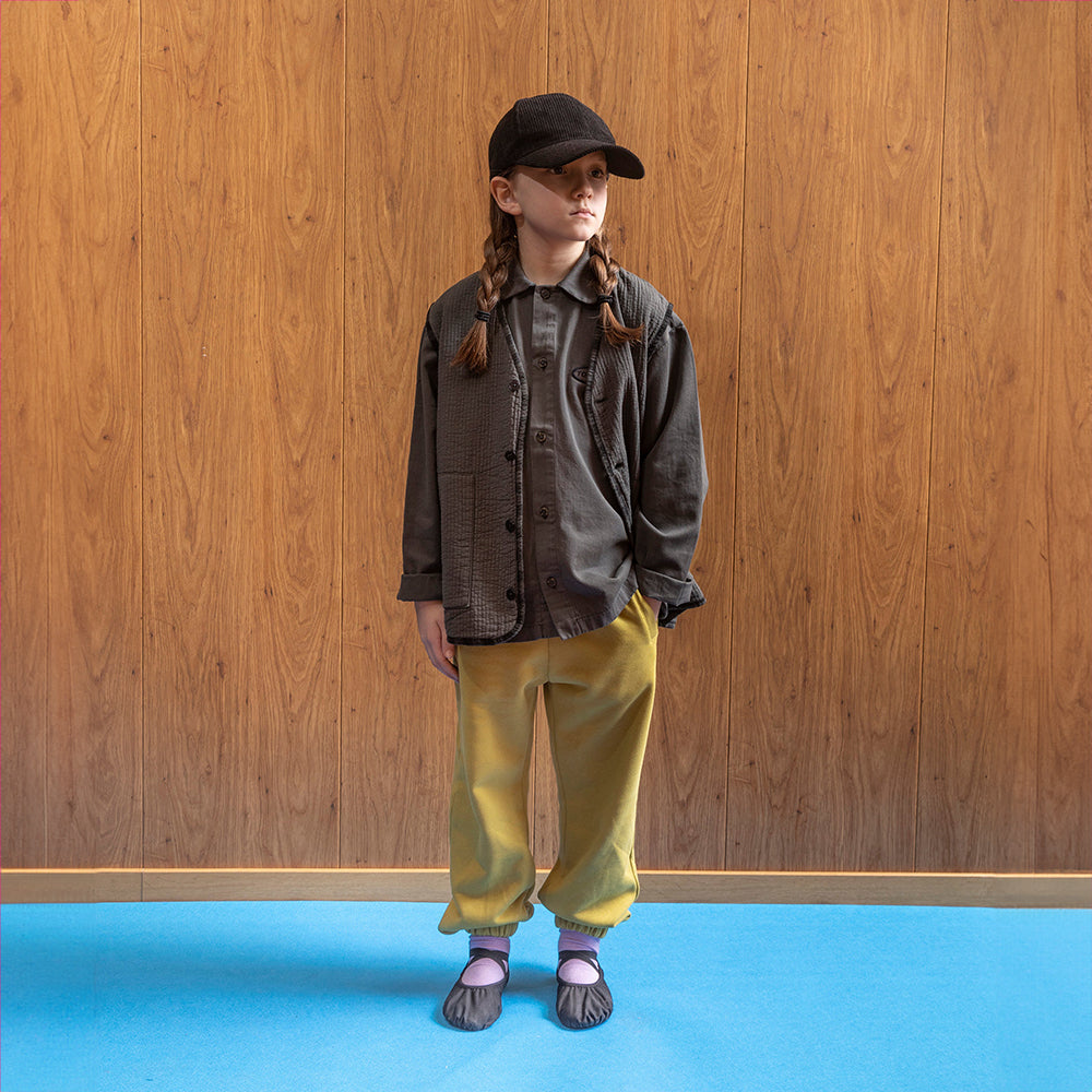 A white girl standing against a brown panelled wall in a room with blue floors wearing a black corduroy hat with two pigtails. She is also wearing a brown jacket with black buttons and underneath that a brown button up shirt. This is paired with acid green cuffed hem joggers, purple socks ad pair of brown ballet pumps. 