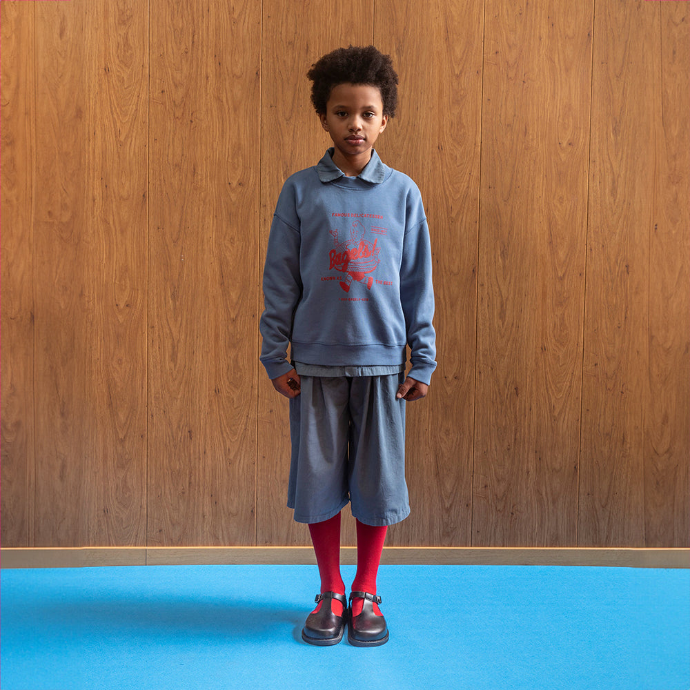 A mixed race boy wears the Tom & Boy blue bagel sweatshirt with the dyed blue short trousers from the same range. He wears them with red over the knee socks and vintage style school shoes. 