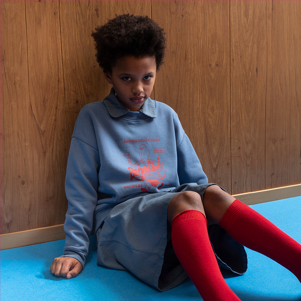 A mixed race boy leans against a wood panelled wall and is wearing the Tom & Boy blue bagel sweatshirt with the dyed blue short trousers from the same range. He wears them with red knee high socks