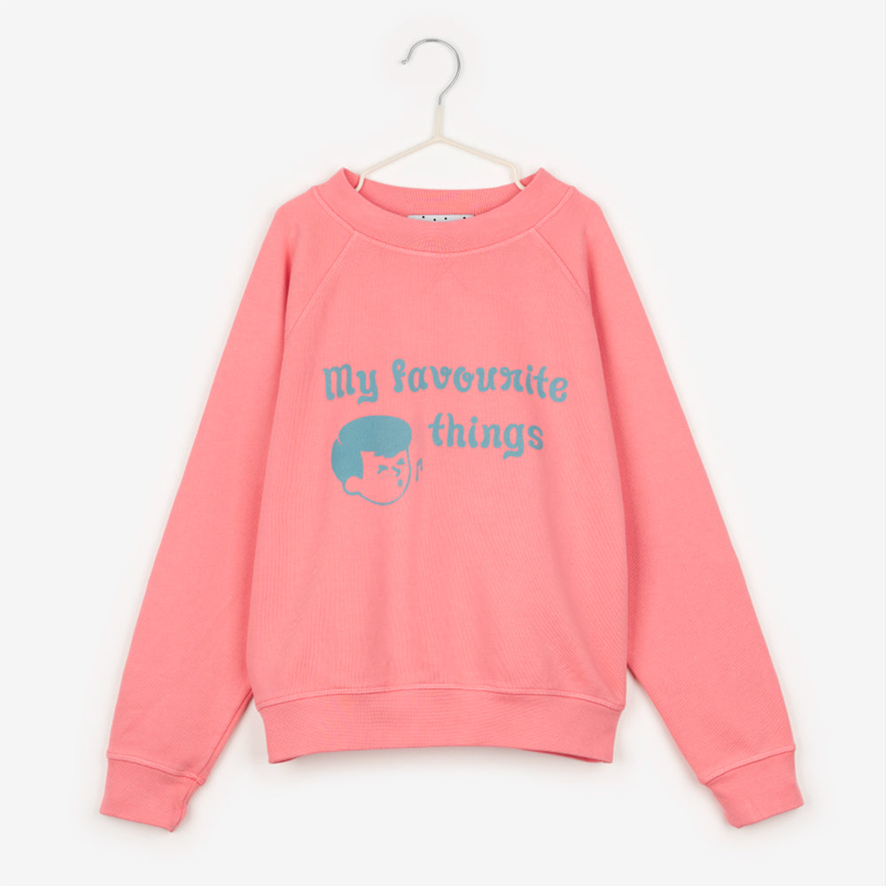 An image of a bright/coral pink sweatshirt hanging up on a white hanger against the background of a white wall. On the sweatshirt in baby blue writing it says 'my favourite things' next to the face of a cartoon boy, who is also drawn in blue, whistling with a music note next to his mouth. 