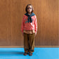 A boy, wearing purple circle frame glasses stands against a wood panelled wall, with blue floors, wearing a bright pink/coral sweatshirt. On the sweatshirt in baby blue writing it says 'my favourite things' next to the face of a cartoon boy, who is also drawn in blue, whistling with a music note next to his mouth. This is paired with a blue scarf around his neck, a white and brown checkered shirt underneath the jumper, a pair of khaki balloon styled trousers paired, both in the collection, & grey clogs. 