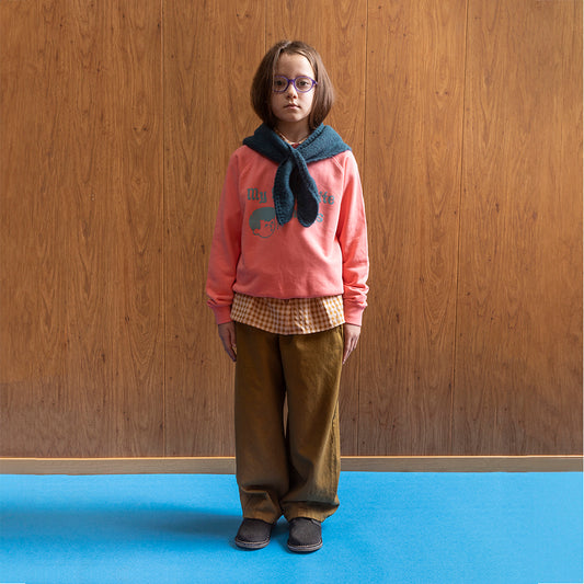 A boy, wearing purple circle frame glasses stands against a wood panelled wall, with blue floors, wearing a bright pink/coral sweatshirt. On the sweatshirt in baby blue writing it says 'my favourite things' next to the face of a cartoon boy, who is also drawn in blue, whistling with a music note next to his mouth. This is paired with a blue scarf around his neck, a white and brown checkered shirt underneath the jumper, a pair of khaki balloon styled trousers paired, both in the collection, & grey clogs. 