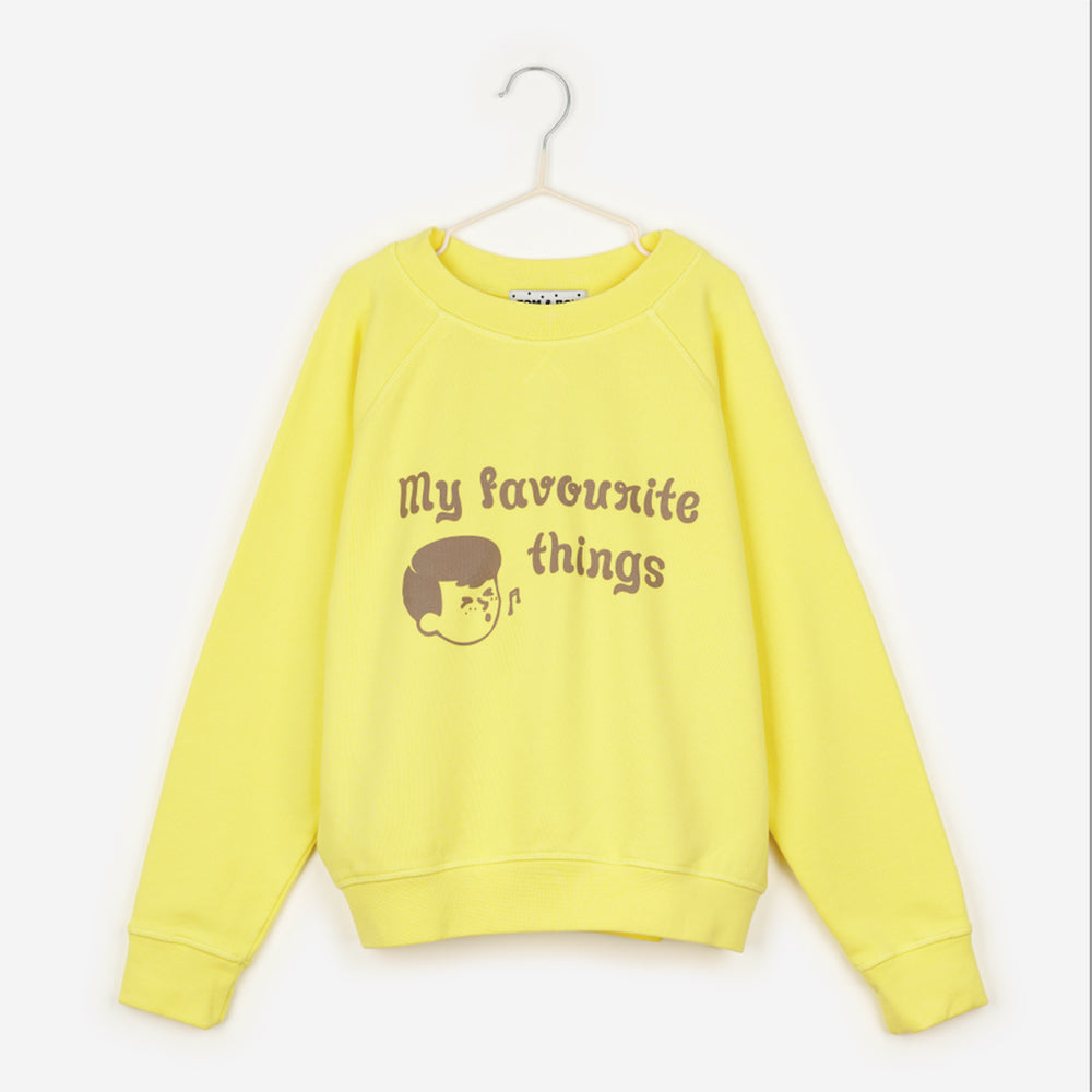 An image of a yellow sweatshirt hanging up on a white hanger against the background of a white wall. On the sweatshirt in brown writing it says 'my favourite things' next to the face of a cartoon boy, who is also drawn in brown, whistling with a music note next to his mouth. 