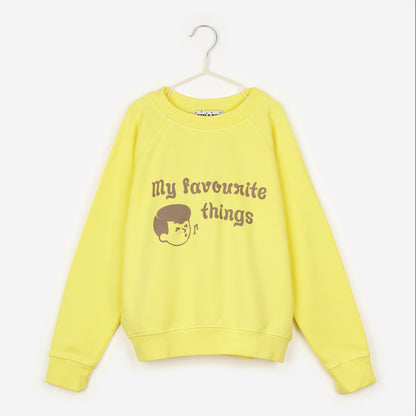 An image of a yellow sweatshirt hanging up on a white hanger against the background of a white wall. On the sweatshirt in brown writing it says 'my favourite things' next to the face of a cartoon boy, who is also drawn in brown, whistling with a music note next to his mouth. 