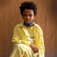 An image of a mixed race boy sitting in front of a brown panelled wall, with his legs crosses, wearing a two piece yellow corduroy trouser and button up shirt set. Underneath the shirt is the yellow Tom&Boy favourite things sweatshirt. He also has a blue bandana around his neck. 