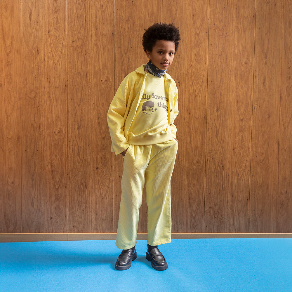 An image of a mixed race boy standing in front of brown panelled wall and a room with blue flooring wears a two piece yellow corduroy trouser and button up shirt set. Underneath the shirt is the yellow Tom&Boy favourite things sweatshirt. He also has a blue bandana around his neck and has on a pair of black loafers. 