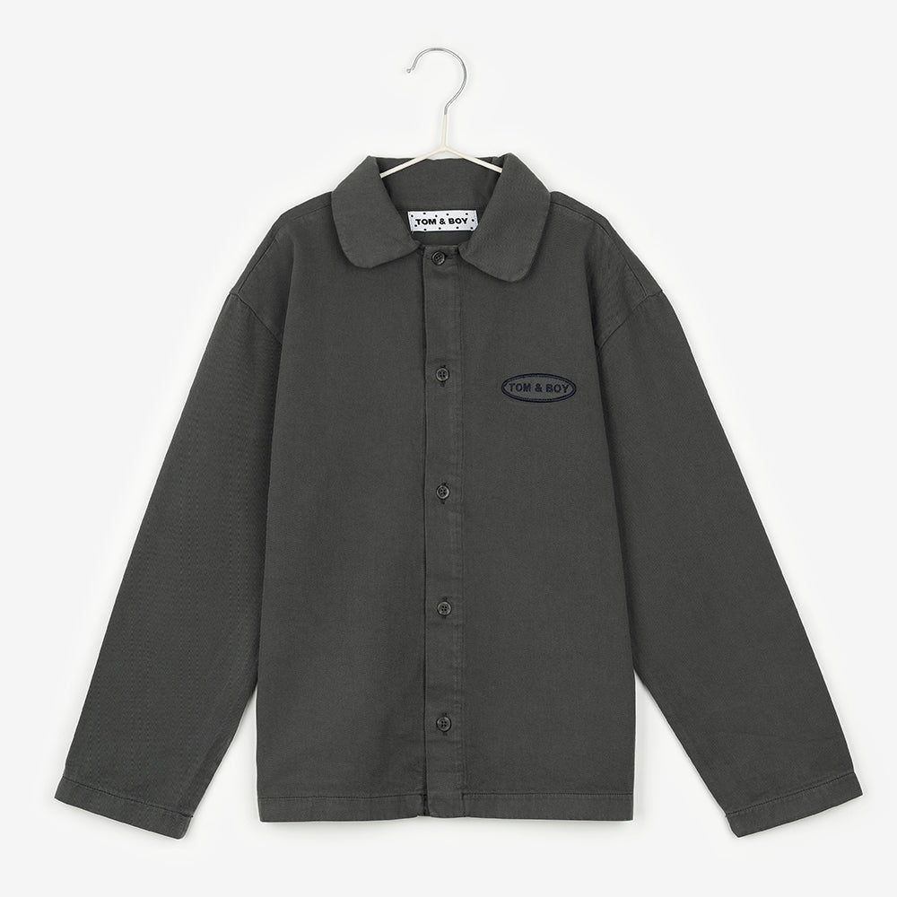 An image of a dark grey button up shirt hanging up on a white hanger against the background of a white wall. On the shirt, to the right hand side of the buttons it says Tom & Boy in black stitch within a black circle, similar to the design of a car mechanics work shirt.