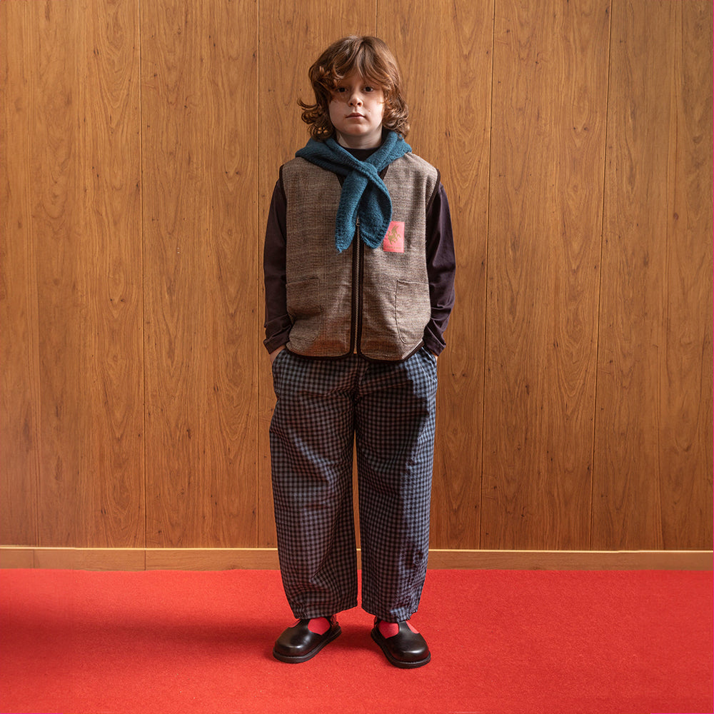 A white boy stands in front of a brown panelled wall in a room with red floors wearing a brown checkered/gingham zip up vest. Underneath the vest is a long sleeve brown shirt. This is paired with blue gingham trousers, red socks with vintage style school shoes and a blue scarf around the neck. 