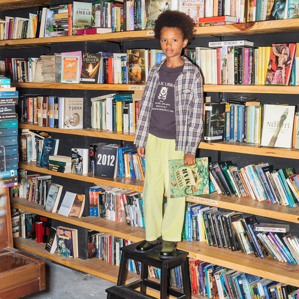 An image of a mixed race by standing on a black stool in a library wearing the brown Tom&Boy checkered over-shirt with a jumper underneath the says public library with the image of a kitten reading. This is paired with yellow trousers and vintage school shoes. He is also holding a book in his hand.