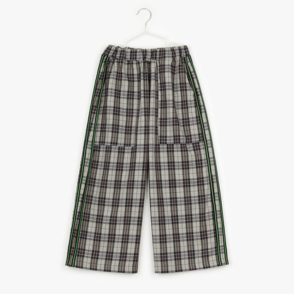 An image of a pair of brown checkered/gingham trousers with 2 green pinstripes, with a black pipe border around it, on either side of the trouser leg hanging up on a white hanger against the background of a white wall. 