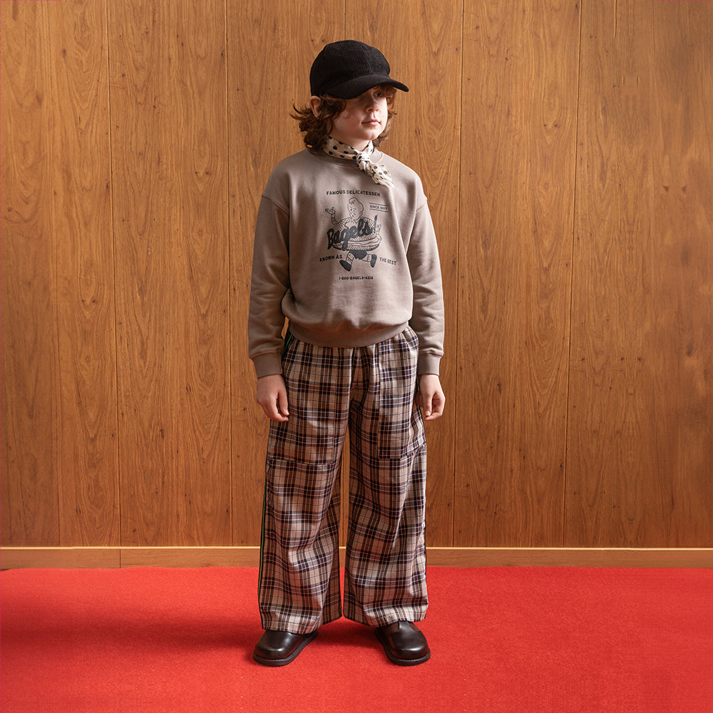 A white boy standing in front of brown panelled wall in a room with red floors wearing a black corduroy hat with a white satin scarf with black dots on it around his neck. He is wearing a jumper that depicting a boy running along with a giant bagel around his waist. It says "famous delicatessen... Bagels!" at the top of the logo and "known as the best" at the bottom, and also "since 1897" and brown checkered/gingham trousers with a pair of vintage style school shoes. 