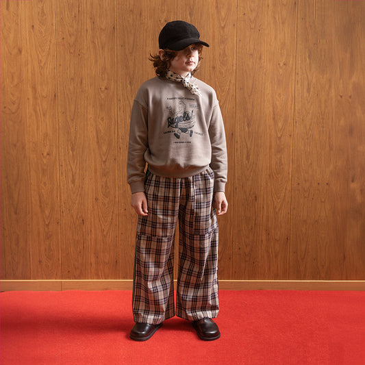 A white boy standing in front of brown panelled wall in a room with red floors wearing a black corduroy hat with a white satin scarf with black dots on it around his neck. He is wearing a jumper that depicting a boy running along with a giant bagel around his waist. It says "famous delicatessen... Bagels!" at the top of the logo and "known as the best" at the bottom, and also "since 1897" and brown checkered/gingham trousers with a pair of vintage style school shoes. 