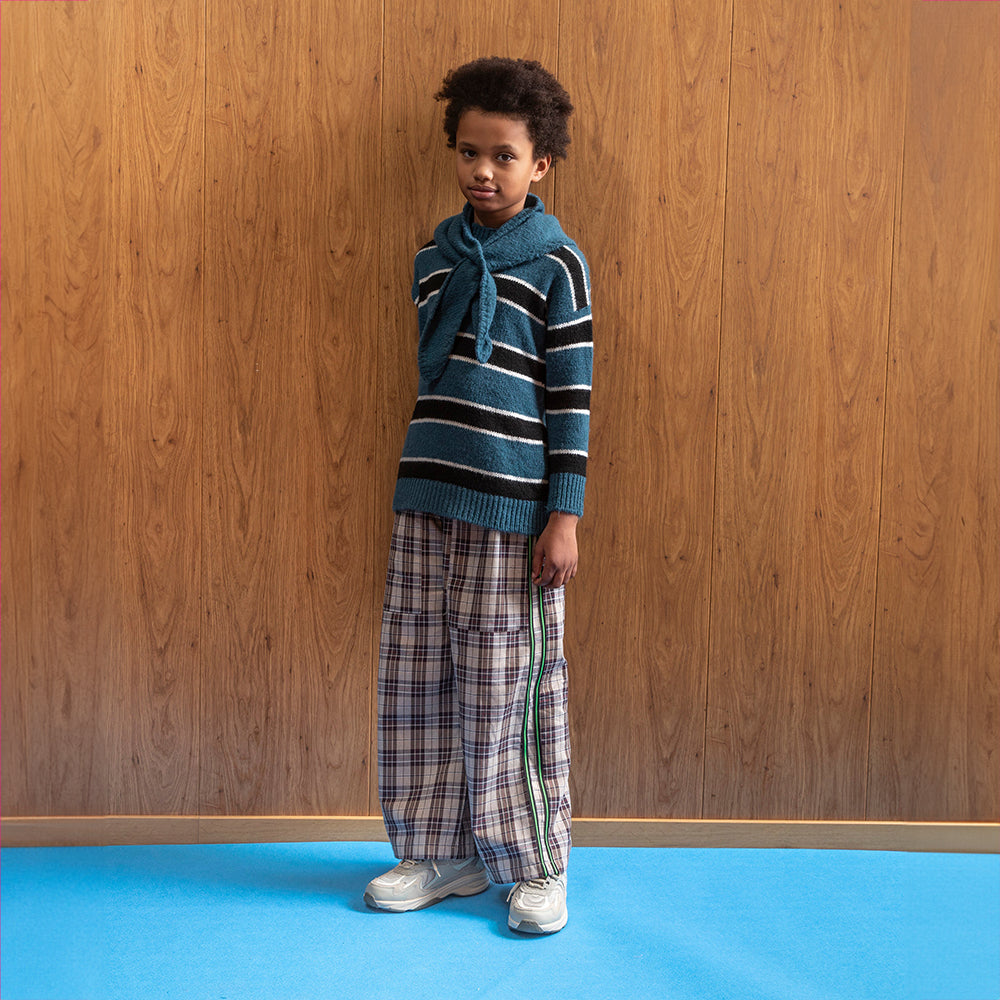 A mixed race boy standing in front of a brown panelled floor in a room with blue flooring wears a blue knitted jumper with thick black stripes that have a white border around them. This is paired with a pair of brown checkered/gingham trousers with 2 green pinstripes, with a black pipe border around it, on either side of the trouser leg. He also has on a blue scarf and a pair of grey trainers. 