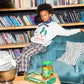 An image of a mixed race boy sitting on the arm of a blue crushed velvet sofa chair in front of a shelf of books wears a white jumper that says Tom&Boy in red and Lower East Side, NY underneath it with two green pickles wearing red boots holding hands. This is paired with f a pair of brown checkered/gingham trousers with 2 green pinstripes, with a black pipe border around it, on either side of the trouser leg and a blue bandana around his neck. In front of him is a table with a jar of pickles & backgammon. 