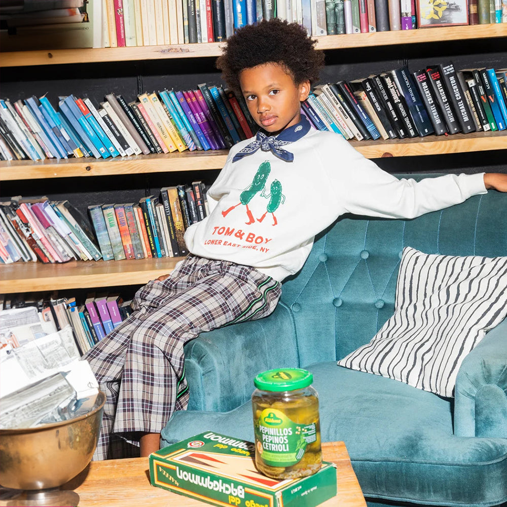 An image of a mixed race boy sitting on the arm of a blue crushed velvet sofa chair in front of a shelf of books wears a white jumper that says Tom&Boy in red and Lower East Side, NY underneath it with two green pickles wearing red boots holding hands. This is paired with f a pair of brown checkered/gingham trousers with 2 green pinstripes, with a black pipe border around it, on either side of the trouser leg and a blue bandana around his neck. In front of him is a table with a jar of pickles & backgammon. 