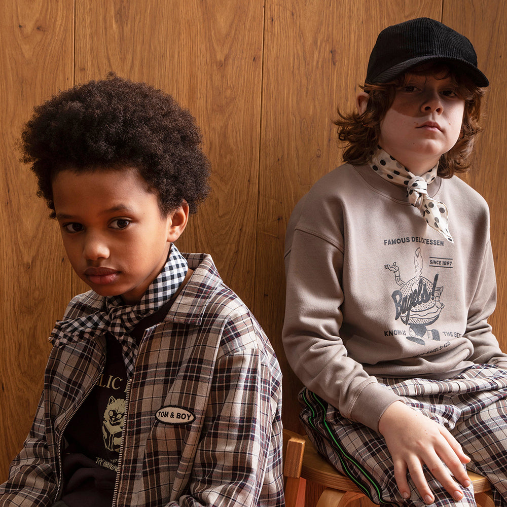 An image of a mixed race boy against a  brown panelled wall, sitting down, wearing the brown Tom&Boy checkered over-shirt with a jumper underneath the says public library with the image of a kitten reading, paired with a bandana around his neck. Next to him is a white boy sitting down with a black corduroy hat on, a white satin scarf with black spots on around his neck, a brown jumper that has a boy running along with a giant bagel around his waist,  saying "famous delicatessen and brown gingham trousers.