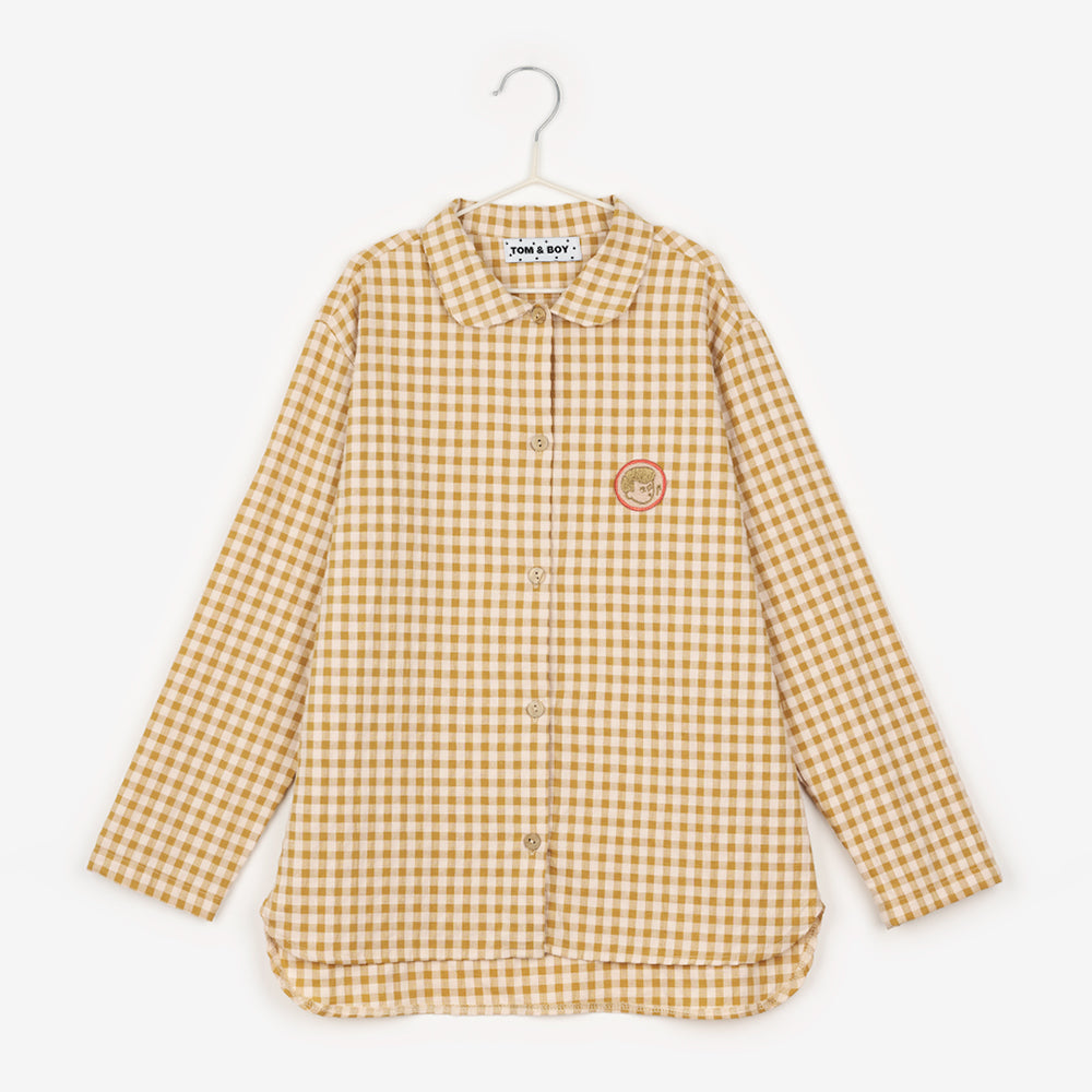 An image of a yellow checkered/gingham button up shirt with an image of boy whistling to the right of the buttons, inside a pink circle, hanging up on a white hanger against the background of a white wall.