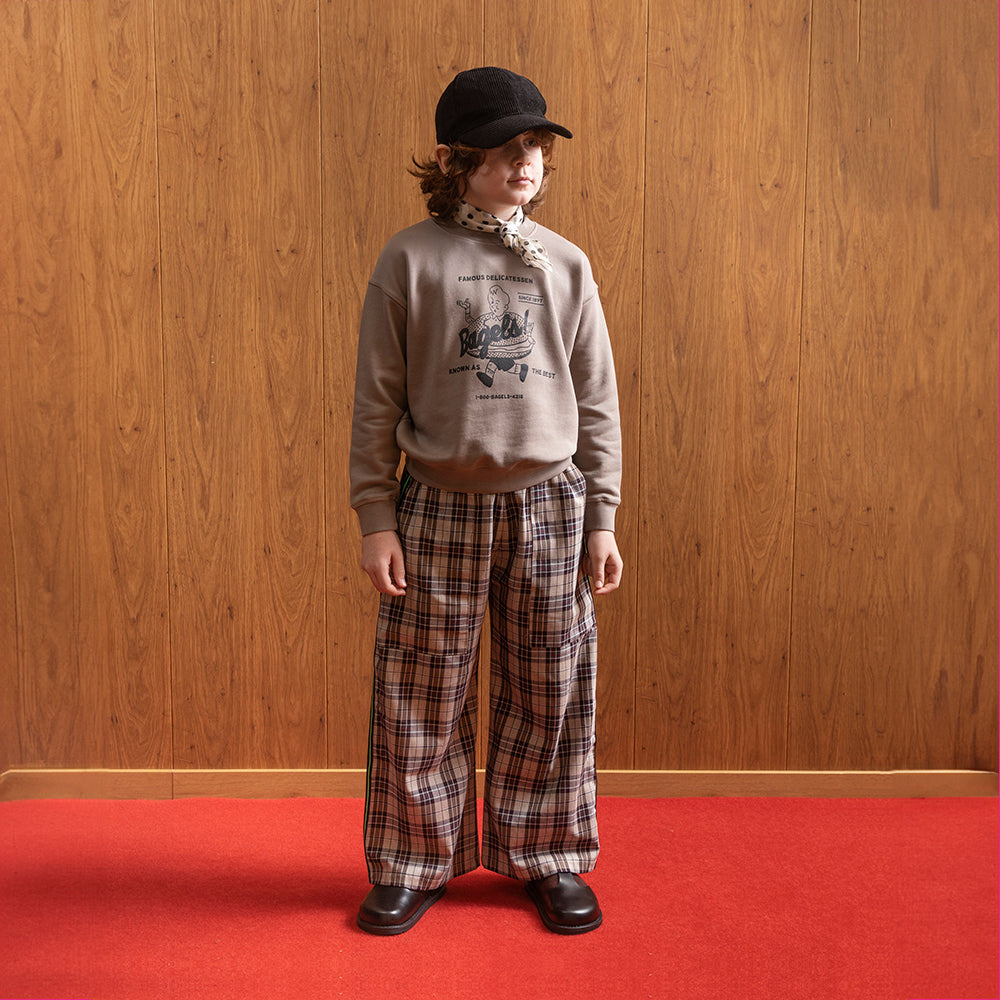 A white boy standing in front of brown panelled wall in a room with red floors wearing a black corduroy hat with a white satin scarf with black dots on it around his neck. He is wearing a jumper that depicting a boy running along with a giant bagel around his waist. It says "famous delicatessen... Bagels!" at the top of the logo and "known as the best" at the bottom, and also "since 1897" and brown checkered/gingham trousers with a pair of vintage style school shoes. 