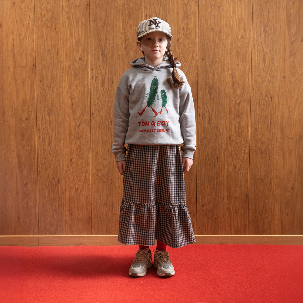 A girl standing in front of a brown panelled wall in a room with red floor wearing a cream hat that says NY on it. This is paired with a grey hoodie with two green pickles smiling, holding hands and walking with red boots on. Below the cartoon pickles it says Tom & Boy in red and below that it says Lower East Side, NY., and a gingham ankle length skirt. She is wearing grey and white trainers. 