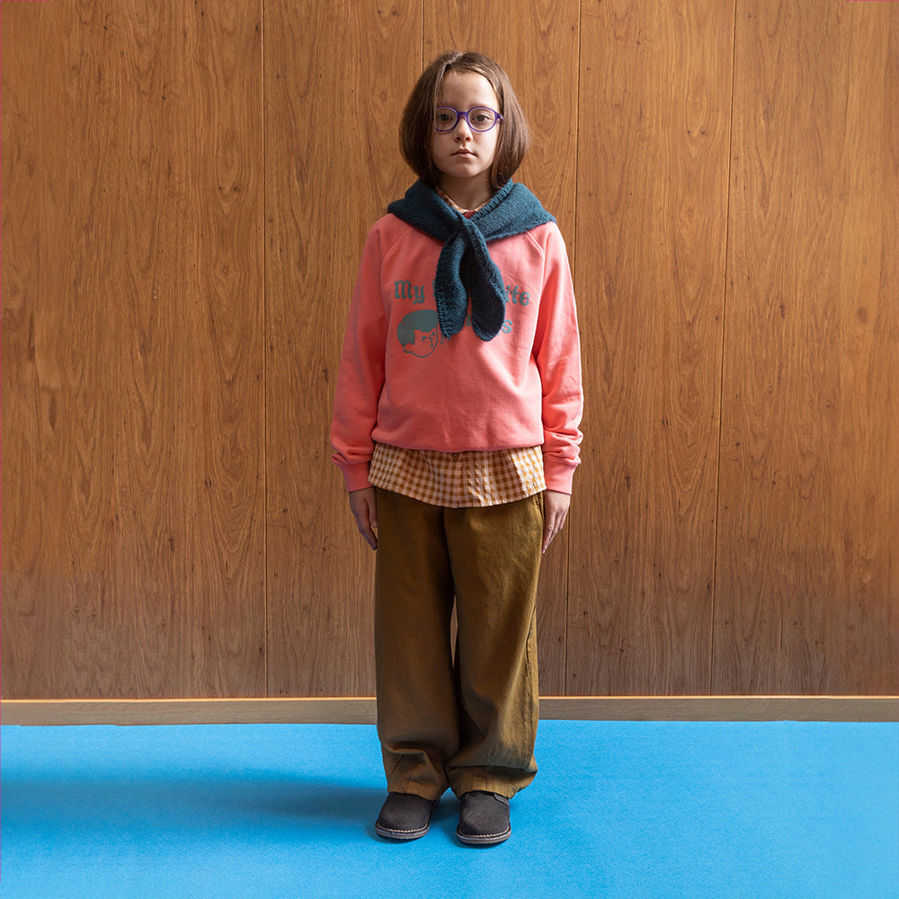 A boy, wearing purple circle frame glasses stands against a wood panelled wall, with blue floors, wearing a bright pink/coral sweatshirt. On the sweatshirt in baby blue writing it says 'my favourite things' next to the face of a cartoon boy, who is also drawn in blue, whistling with a music note next to his mouth. This is paired with a blue scarf around his neck, a white and brown checkered shirt underneath the jumper, a pair of khaki balloon styled trousers paired, both in the collection, & grey clogs. 