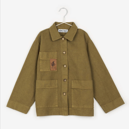 A khaki workwear boxy jacket that has brown wooden buttons going down the middle and two square pockets on the bottom, either side. On the left hand side of the jacket is brown donkey patch on another square pocket that has Tom & Boy beneath the donkey. 