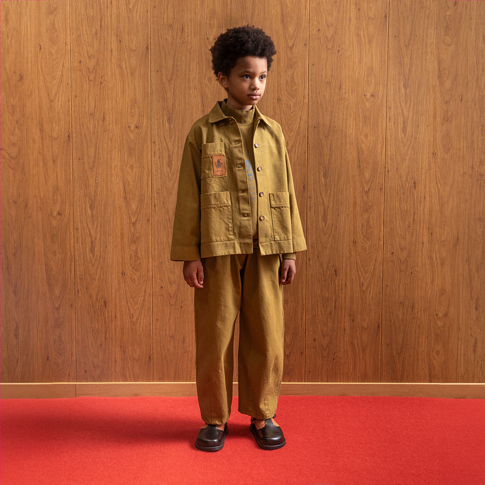 A boy standing in front of a brown panelled wall in a room with red floors wears a khaki workwear boxy jacket. The jacket has brown buttons and two square pockets on the bottom, either side. On the left hand side of the jacket is brown donkey patch on another square pocket. This is paired with of khaki balloon leg trousers and a pair of black vintage school shoes and orange socks. 