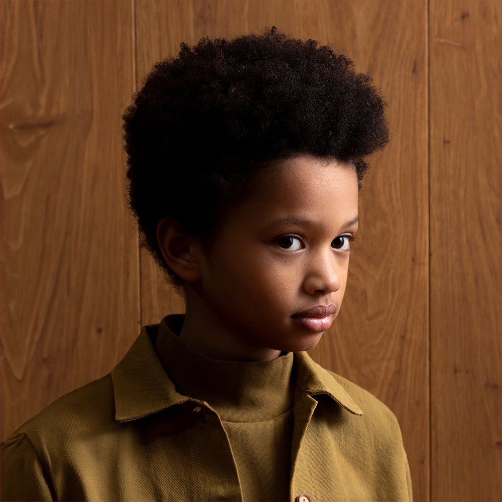 A closeup headshot of a mixed race boy standing in front of a brown panelled wall. He is wearing a high neck khaki jumper and a khaki jacket which appears to have wooden buttons going down the front. 