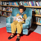 A boy sits in a blue crushed velvet chair in front of shelves of books reading a manga book. He is wearing a khaki workwear boxy jacket and khaki balloon leg trousers. Around his neck is a blue knitted jumper that has black stripes with a white border around th outside of them. This is paired with black vintage style school shoes and orange socks . 