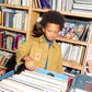 A boy looking at music records in front of shelves of books. He is wearing a high neck blue jumper and on top of that a a khaki workwear boxy jacket. The jacket has brown buttons and two square pockets on the bottom, either side. On the left hand side of the jacket is brown donkey patch on another square pocket.