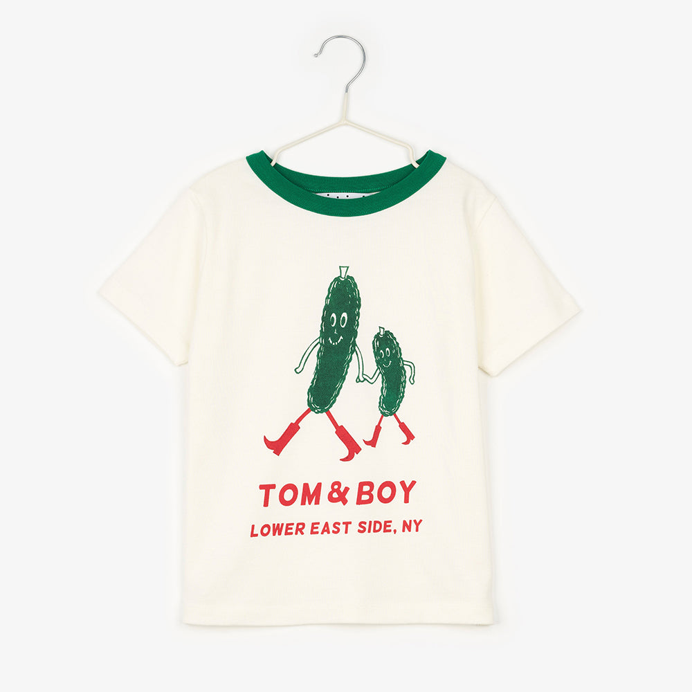 A cream/white short sleeve -shirt with a green neckline. In the middle of the t-shirt there are two green pickles smiling, holding hands and walking with red boots on. Below the cartoon pickles it says Tom & Boy in red and below that it says Lower East Side, NY. 