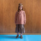 A girl standing in front of a brown panelled wall in a room with blue floors wearing purple glasses and a brown and white knitted hat. This is paired with a dusty pink high neck jumper that has the words public library in dark purple. Beneath that is an image of a kitten reading a book, followed with the opening times of the library and the location and an ankle length gingham skirt. She is also wearing purple socks with brown ankle length suede boots with cream laces. 