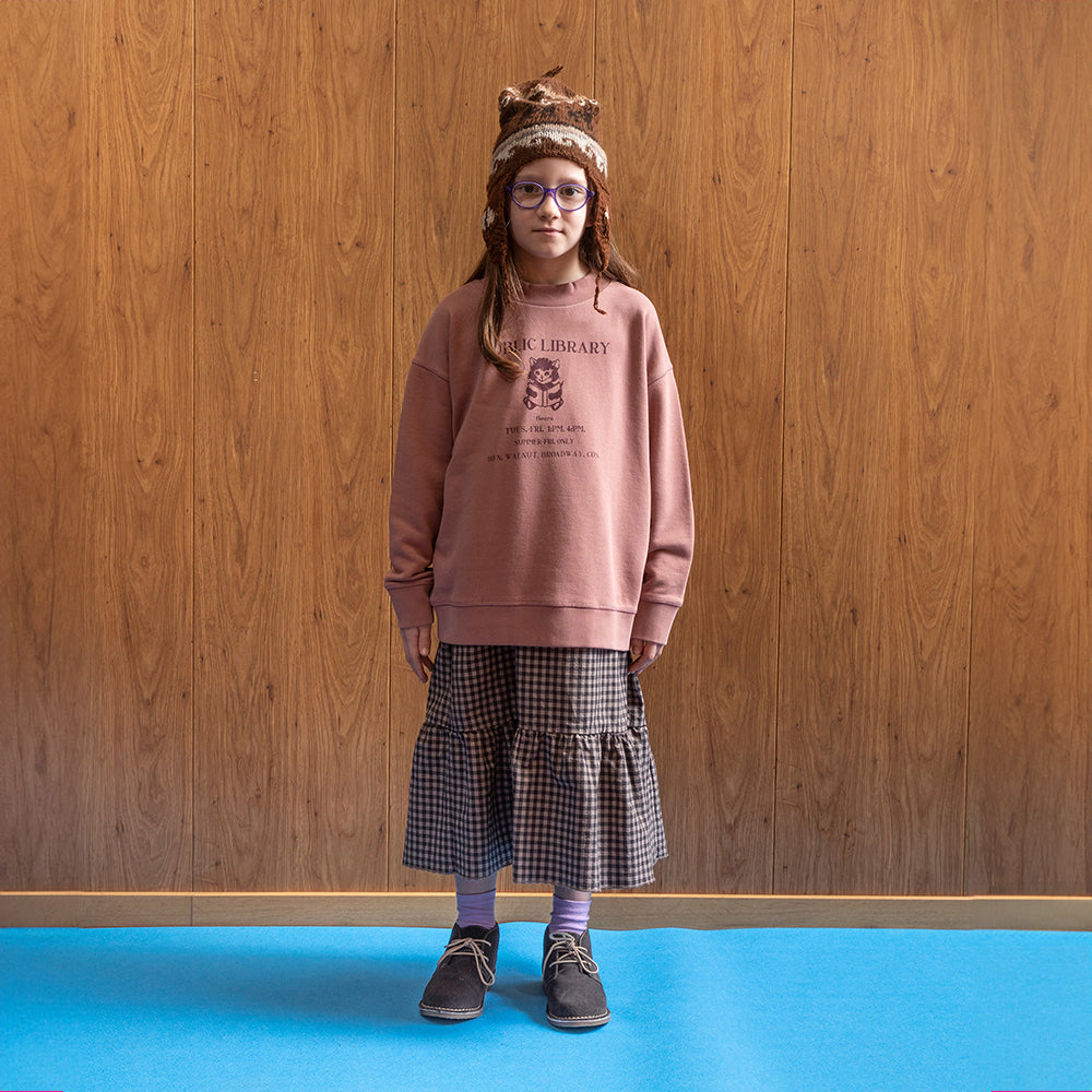 A girl standing in front of a brown panelled wall in a room with blue floors wearing purple glasses and a brown and white knitted hat. This is paired with a dusty pink high neck jumper that has the words public library in dark purple. Beneath that is an image of a kitten reading a book, followed with the opening times of the library and the location and an ankle length gingham skirt. She is also wearing purple socks with brown ankle length suede boots with cream laces. 